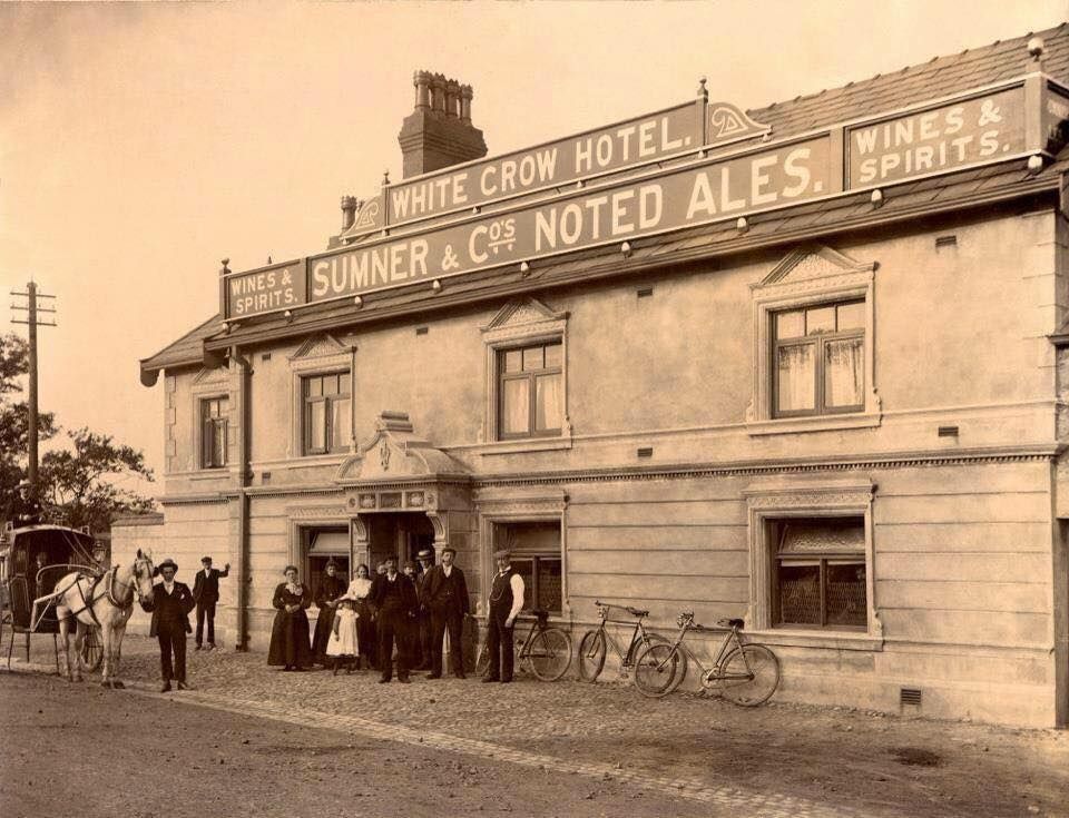 An old sepia photograph of the White Crow Hotel, signwritten Sumner and Co's Noted Ales, Wines and Spirits, with people, a horse and bicycles outside